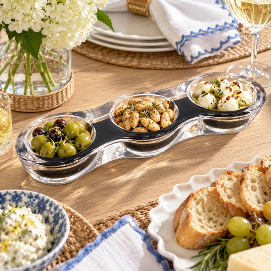 Slate modern acrylic grazing bowl with three clear sections holding olives, herbed almonds and marinated mozzarella on a wood dinner table with bread and grapes