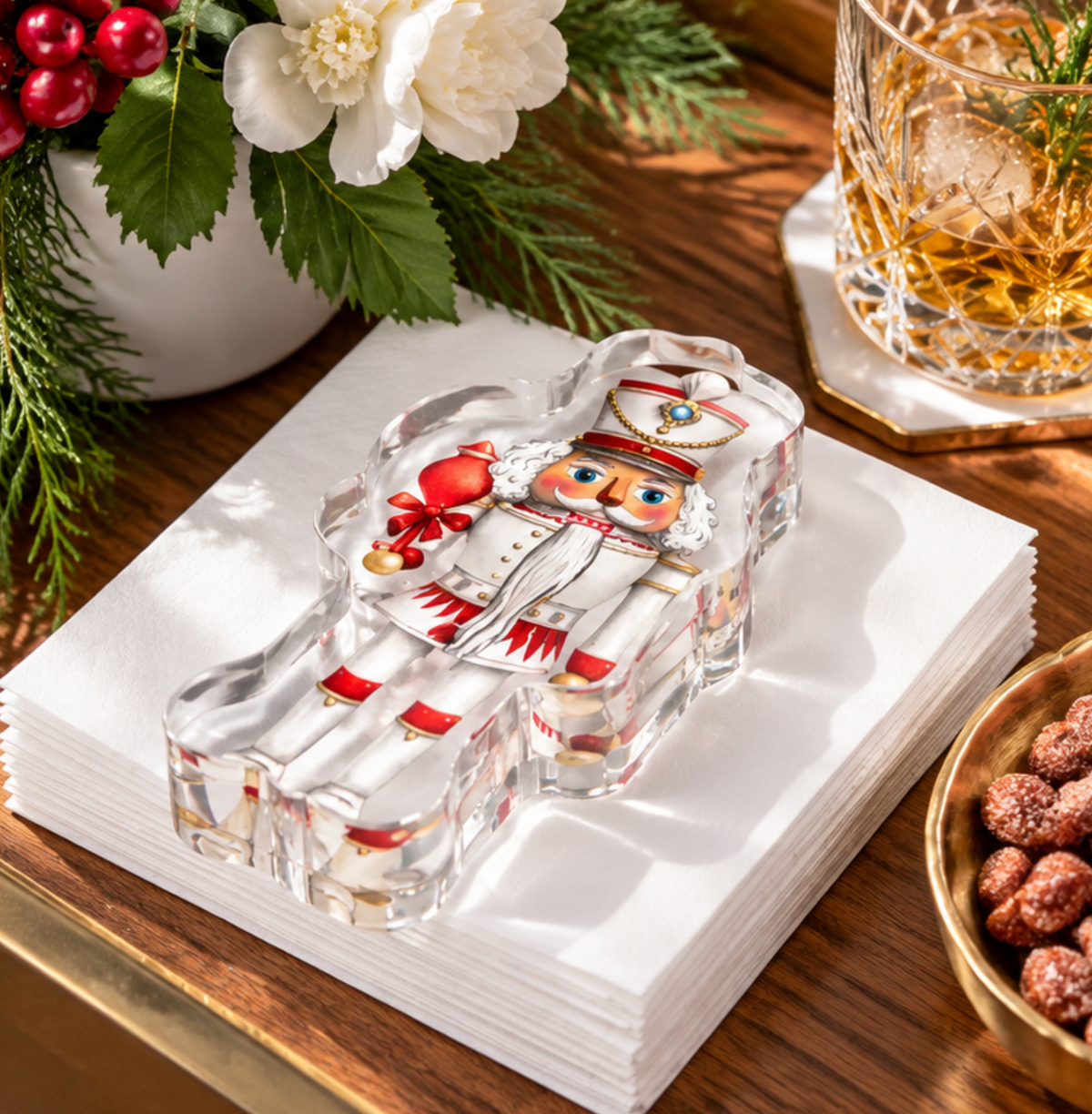 Nutcracker acrylic napkin weight on white cocktail napkins on a wooden table, with white flowers, a crystal glass and a dish of nuts
