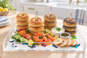 Bagels stacked on the clear acrylic dowels over a white butterfly print base with smoked salmon, cucumber, capers, cream cheese and toast in a sunny kitchen