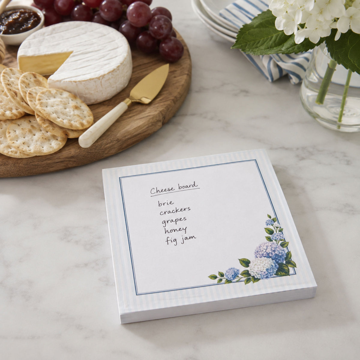 Hydrangea notepad with a handwritten cheese board list beside brie, crackers and grapes