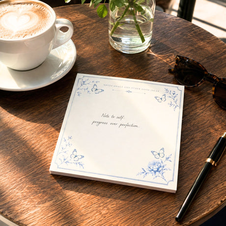 Notes and Goals notepad reading progress over perfection on a wood cafe table