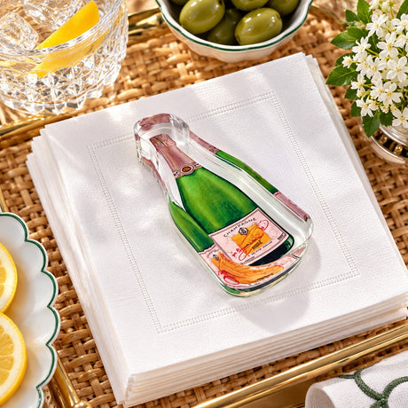 Bubbles Champagne acrylic napkin weight shaped like a green champagne bottle on white cocktail napkins on a rattan tray with olives and lemons