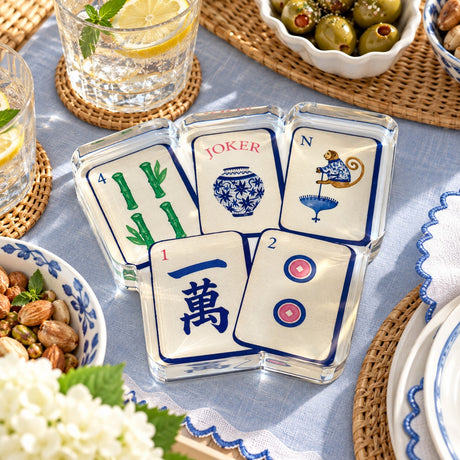 Preppy Mahjong acrylic napkin weight with 4 Bam, Joker ginger jar, North monkey, 1 Crak and 2 Dot tiles on a pale blue linen tablecloth with olives, nuts and hydrangeas