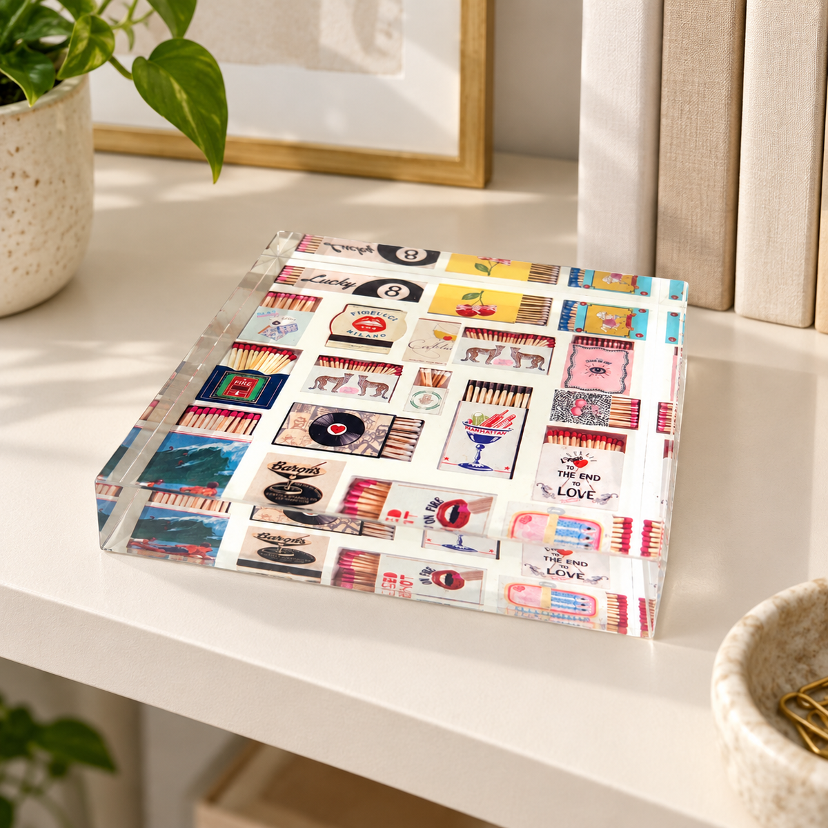 Matchbox acrylic napkin weight lying on a white shelf with books and a potted plant, showing the vintage-style matchbox collage