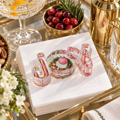 JOY acrylic napkin weight on white cocktail napkins on a gold bar cart with a champagne coupe, cranberries and rosemary