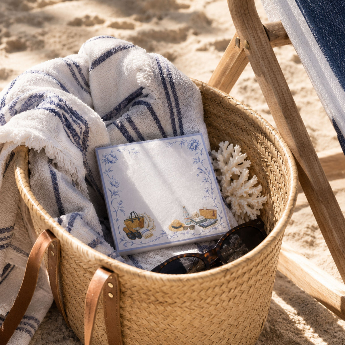 Proper Beach Club notepad in a straw beach basket with a striped towel and coral