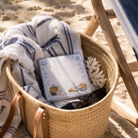 Proper Beach Club notepad in a straw beach basket with a striped towel and coral