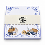 Beach notepad in its MH band with a lavender border, blue florals, a straw tote, sun hat and sandals