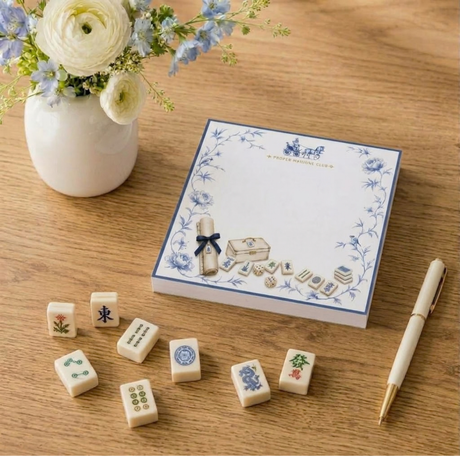 Proper Mahjong Club notepad with horse carriage and blue peony border beside mahjong tiles, a vase of ranunculus and a pen