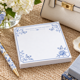 Notes, Goals and Other Good Ideas 5x5 notepad with blue butterflies and peonies on a woven table beside a cream handbag, hydrangeas and a blue-and-white pen