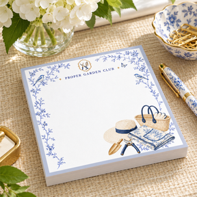 Proper Garden Club 5x5 notepad with blue toile vines, garden tote, sun hat and pruners on a woven table with hydrangeas and a gold pen
