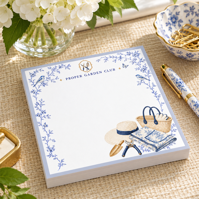 Proper Garden Club 5x5 notepad with blue toile vines, garden tote, sun hat and pruners on a woven table with hydrangeas and a gold pen